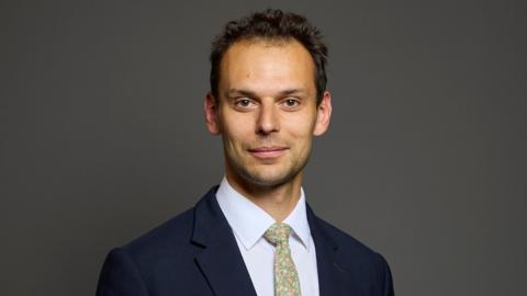 Josh Simons has dark hair, a pale green tie, white shirt and blue suit