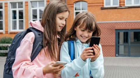 Two young girls outside a school building, both looking at mobile phones. Both are wearing backpacks and have long brown hair. The girl on the left is wearing a pink jacket, while the other girl is wearing a black jacket.