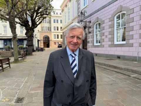Deputy Sir Philip Bailhache is pictured outside the States Assembly. He is wearing a grey coat and a blue shirt. 
