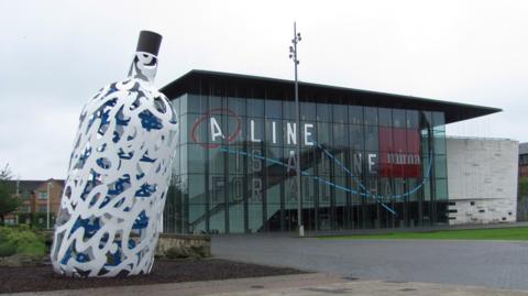 A general view of MIMA in Middlesbrough. The building is made of glass and has a flat roof. There is a large sculpture of a white and blue bottle to one side.