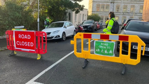 A woman in a yellow high visibility jacket stands next to two plastic barriers. One is red and says 'Road Closed', the other is yellow and has a green sign indicating the road is open to pedestrians, wheelchair users, cyclists and those on scooters.