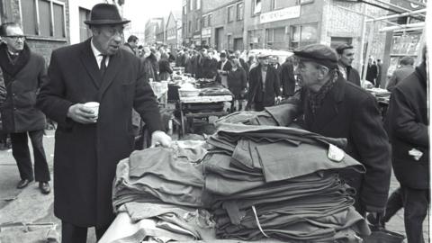 Black and white image of two men in hats and overcoats at an outdoor market. They are chatting over a pile of trousers on a market stall.