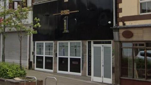 Bar 1 on Front Street in Consett. The bar has a black frontage and its name appears in gold lettering. There are six windows to the front of the building with a plants in a planter across the pavement.