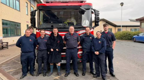 Seven men and one woman standing in front of a red fire engine wearing matching black trousers, black shoes and blue jumpers or shirts