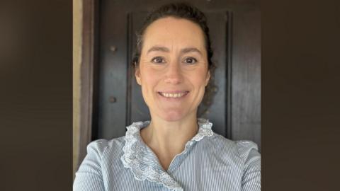 A woman smiling at the camera. She is wearing a blue and white striped top with white lace. Her brown curly hair is tied back. Behind her is a brown background. 
