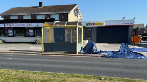 A yellow framed bus stop outside a Go Local Extra shop and The New Johnny Todd pub in Kirkby is cordoned off by police. Blue plastic sheeting can be seen and the shop's shutters are down.