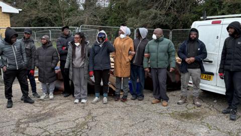 A group of people standing in front of a fence and a white van. They are wearing winter coats and not all of them are looking at the camera.