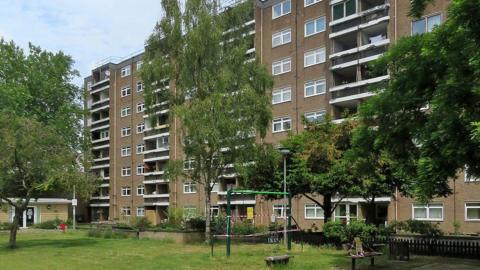 Rectangular building with uniformly spaced double windows and columns of balconies. In front of it are trees and outdoor play equipment. 