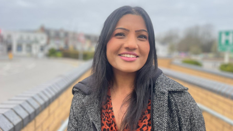 A woman with long black hair is wearing a white and black coat with orange and black underneath. Buildings in the distance are blurred.