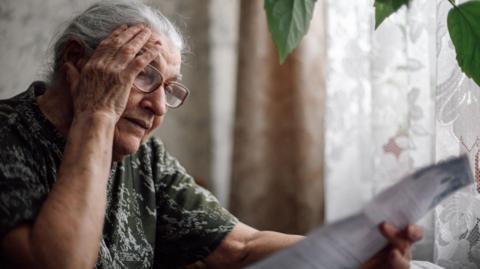 Older woman looks at a bill with curtains closed behind her.