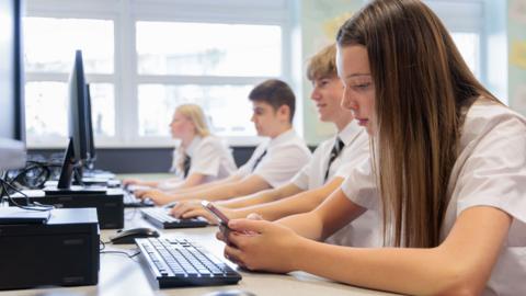 Female high school student looking a smartphone in a school IT class while sitting at a computer