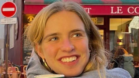 Clarissa Nicholls, who has light hair and is wearing a grey top, smiling while outside a coffee shop in Paris