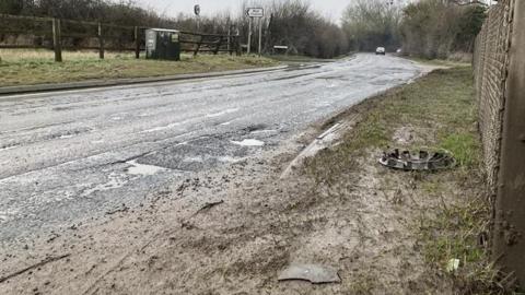 A main road, which is pockmarked with ridges and craters. There is some debris on the side of the road, including a hubcap. It is a wet day so the holes are puddles.