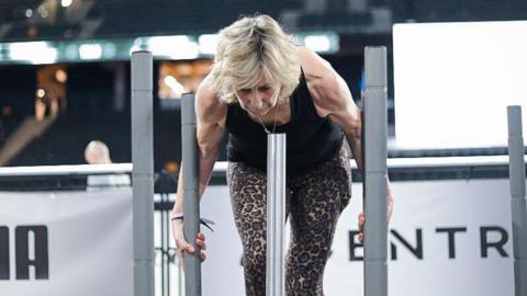 A woman pushes a weighted sled during a Hyrox competition. She has short blonde hair and is wearing a black gym tank top and animal print leggings.