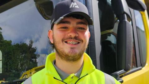 Daniel Kinsella has a dark beard and is wearing a black baseball cap and yellow high-vis jacket. He is standing in front of a yellow bin lorry and smiling for the camera.