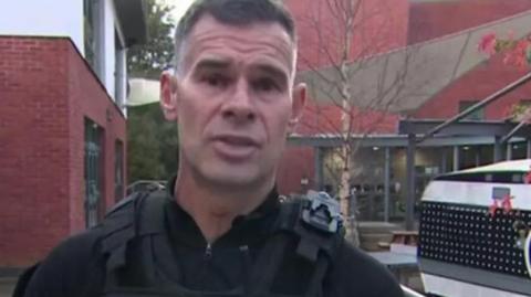 Harry Tangye has short hair and is clean-shaven. He is wearing a black police uniform and is looking straight at the camera