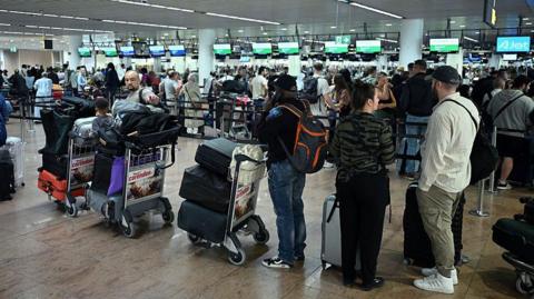 Queues of people with trolleys filled with baggage wait at a busy airport terminal 