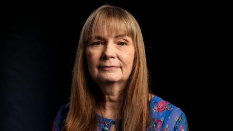 Jo Berry, a woman with long brown hair and a blue top with pink flowers on it