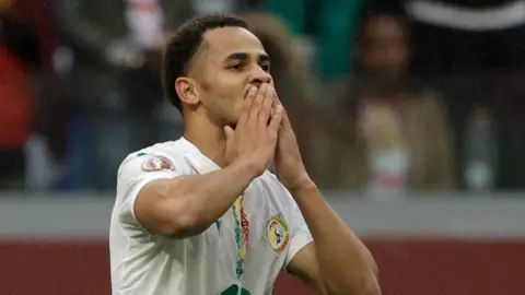 Iliman Ndiaye, wearing a white Senegal shirt with green trim, holds his hands to his mouth as he celebrates a goal
