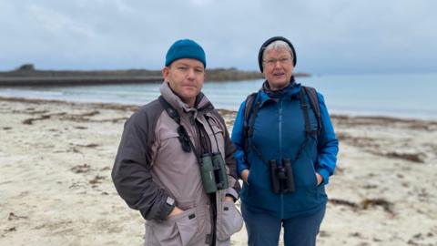 A man in a blue hat and a grey jacket stands on a beach with binoculars next to a woman in a grey hat and a blue jacket, also with binoculars.