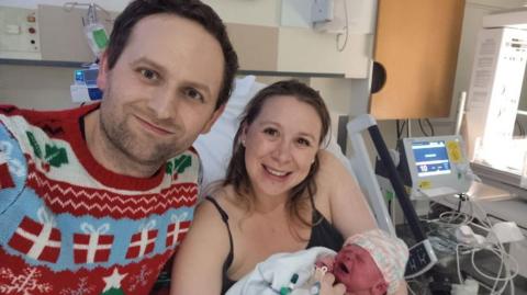 A mum and dad in a hospital ward smiling, with mum holding her newborn baby. The man is wearing a red Christmas jumper, with hospital machinery next to the bed. 