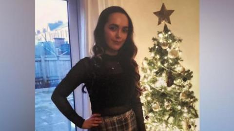 Mia is wearing a tartan skirt and black jumper and is stood in a house in front of a Christmas tree.