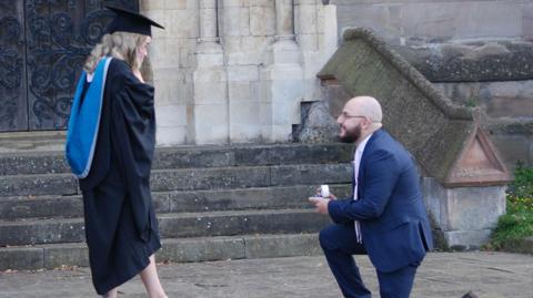 A woman in a  blue and black graduation cloak and black hat. She has blonde hair and is holding her hands on her face in shock.
A man with shaved head, glasses and a brown beard is down on one knee holding a ring box. He is wearing a navy blue suit. 