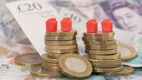 Banknotes and piles of £1 coins with red monopoly houses balanced on top.