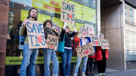 Six people holding placards reading Save T103, except the 0 is an heart. They are all leaning against the wall of a large building.