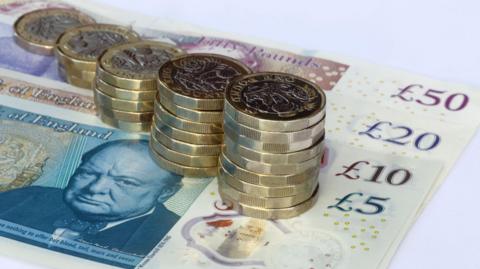 Piles of pound coins which get gradually higher the nearer they are to the camera. The coins are sitting on top of £50, £20, £10 and £5 notes.