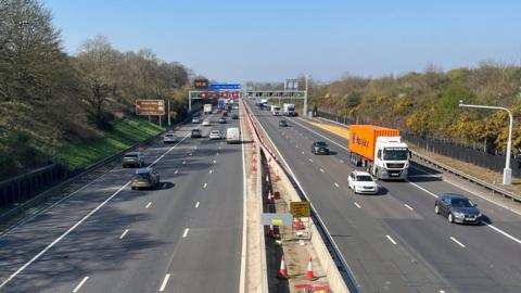 The M1 near junction 13 at Brogborough. There is a brown sign to Woburn Abbey on the left with grass verges and trees on either side of the carriageways. Traffic cones have been moved off the road and have been placed in the central reservation, which has concrete barriers.