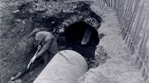 A black and white photo shows a worker digging in a trench beside a large cylindrical pipe, with a small tunnel opening in the exposed earth and a fence running along the edge.