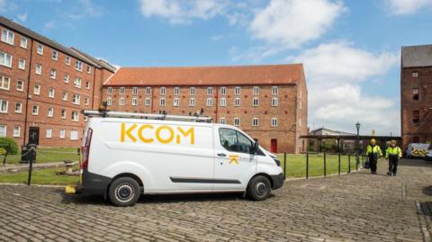 A white van with the letters KCOM in yellow printed on its right-hand side, which is facing the camera, is parked on a cobbled street. Behind it is a block of flats with small windows. Two people wearing yellow headgear and hi-vis jackets, with dark trousers, walk towards the front of the van.