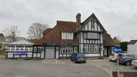 A pub. The building is grey with black external beams. The roof is brown. There is a large car park in front of the pub with four cars in it. The sky is grey