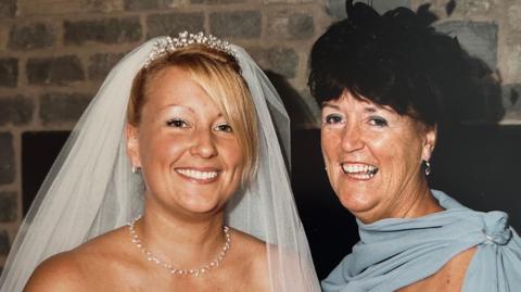 A blonde woman in a white wedding dress stands next to a woman smiling in a blue dress. 