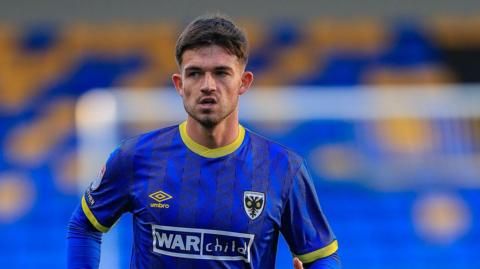 James Furlong jogging on the pitch wearing a blue AFC Wimbledon kit