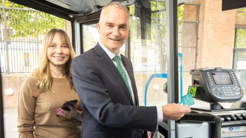 Man in a suit using a credit card to pay onto a bus. Behind him a woman stands smiling holding her phone