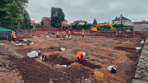 A group of archaeologists working at the site. There are excavators in the background and a member of staff in a high-vis jacket and helmet is working with hand tools in the foreground.