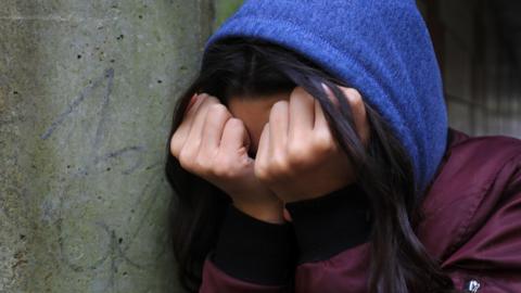 A teenage girl obscures her face with balled fists pressed against her eyes as she leans against a wall