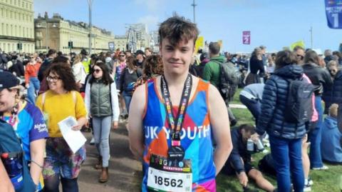 Billy Nunn, a young man wearing a bright blue and orange running vest, looks at the camera. He wears a Brighton Marathon finisher's medal around his neck. A crowd of people are standing and walking behind him. The sky behind him is blue.