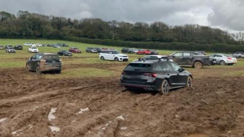 A field covered in wet mud with dep track marks from car tyres. Some of the cars in the foreground are covered in mud while others are parked in the distance.