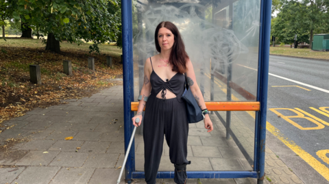A woman wearing a black shirt with thin straps and trousers stands outside a bus shelter on a pavement. She is holding a crutch with one hand. There is a bus lane to the right of the shelter and a park to the left.
