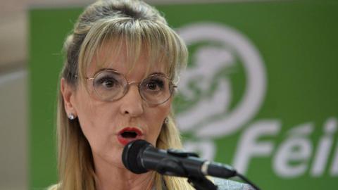 A woman wearing round glasses talks into a microphone. She has shoulder length blonde hair. In the blurred background there is a green poster that says Sinn Fein.