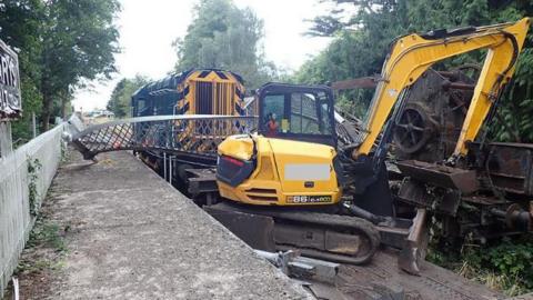 A yellow and black train engine and yellow and digger sit on railway tracks. A wrought iron structure lies between them. A railway platform with steel railings is visible. 