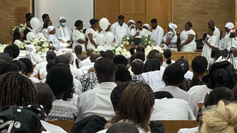 A large group of people dressed predominantly in white are gathered in a spacious hall. The audience is seated on wooden chairs, facing a stage at the front where individuals, also in white, are standing or seated around floral arrangements. The hall has brick walls, high ceilings, and large windows letting in natural light.
