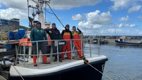 Four fishermen are standing on the deck of a fishing boat in a harbour. Two are men in their forties and two are teenagers. The two youngest are in waterproof gear