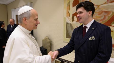 The Pope, wearing white shakes hands with Harry Clark, who is wearing a black suit. 