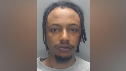 Mugshot of Mohammed. He has black dreadlocks, a thin black moustache and wispy black beard on his chin.
