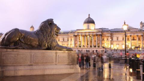 The frontage of the National Gallery photographed from Trafalgar Square with one of the sculptures of a lion at the base of Nelson's Column in the foreground of the image. 