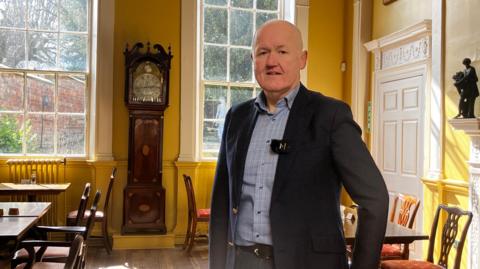 A man in a dark suit jacket and open-necked, checked, grey shirt stands in a large dining room in a Georgian building. Behind him are two large arch windows and a grandfather clock. The sun is shining through the window 
